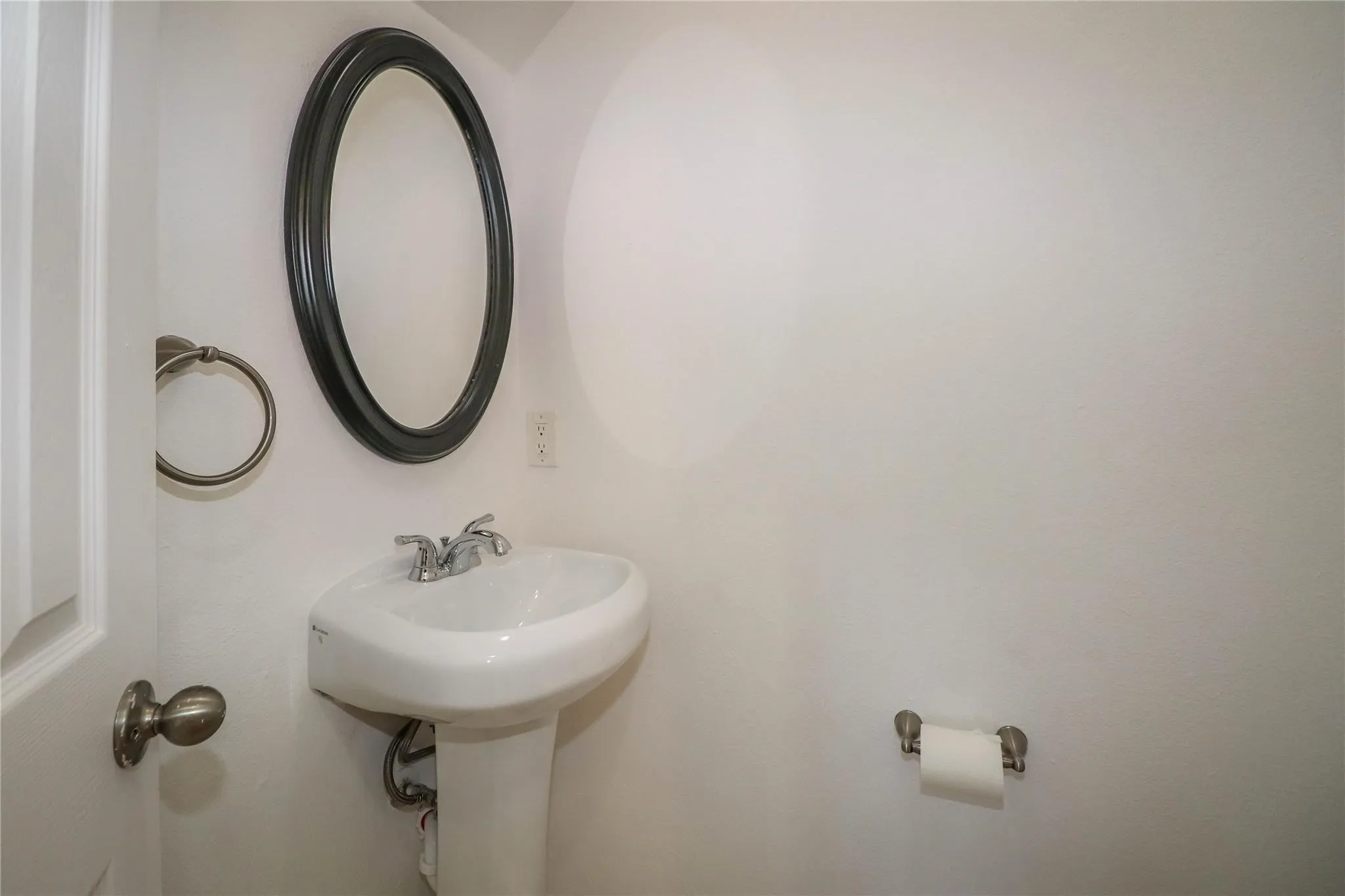 Wall-mounted oval mirror with a dark frame, white pedestal sink, dual-handle chrome faucet, and circular metal towel ring