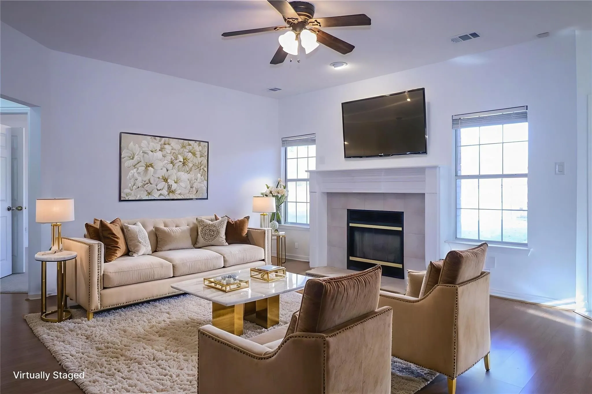 Living area featuring a fireplace with a white mantel and tile surround, large windows, wood-finish flooring, and a ceiling fan with integrated lighting