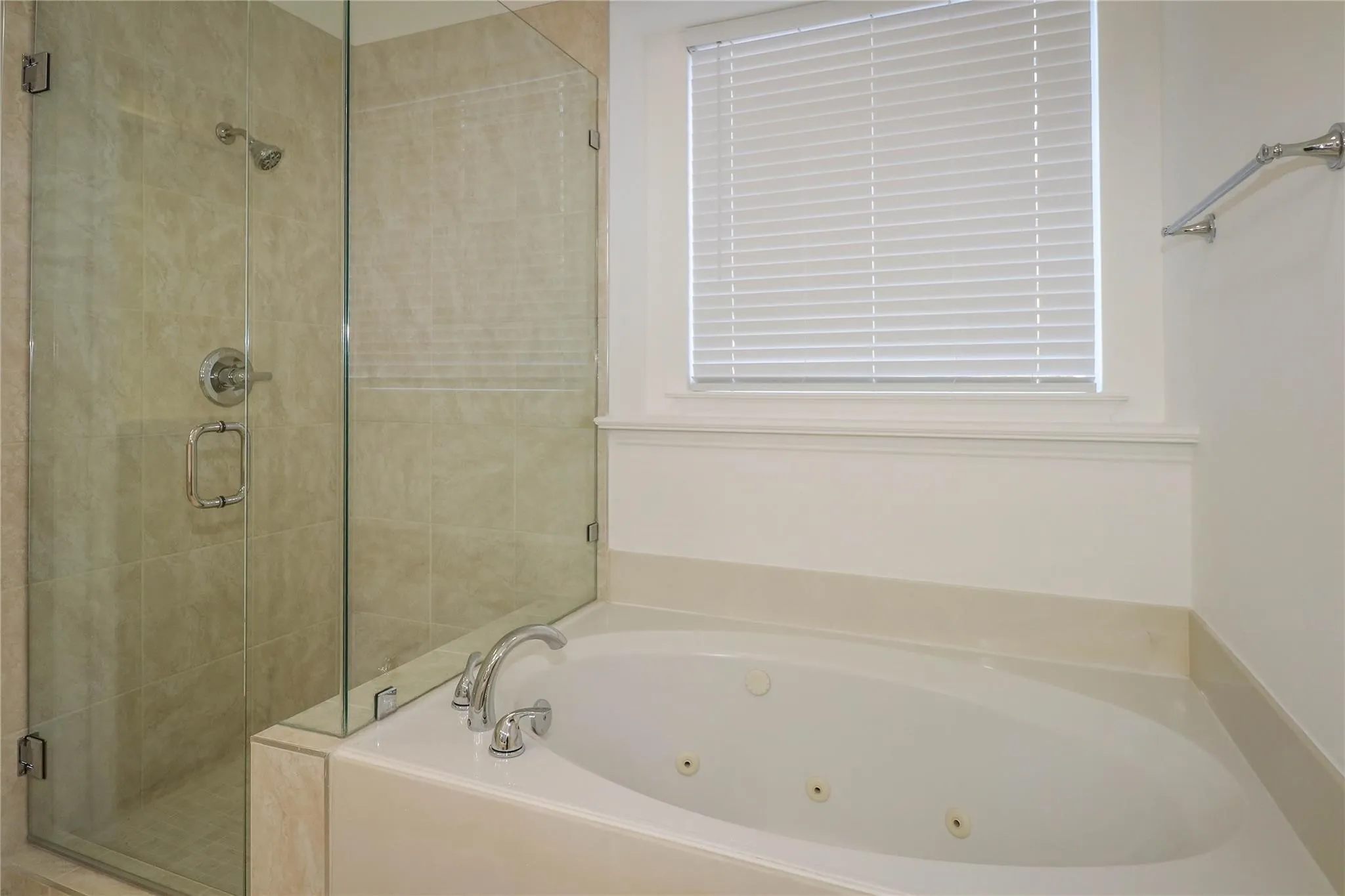 Oval jetted soaking tub with a deck-mounted chrome faucet, separate walk-in shower featuring glass enclosure walls and neutral-toned tile surround, and a recessed window with fitted blinds