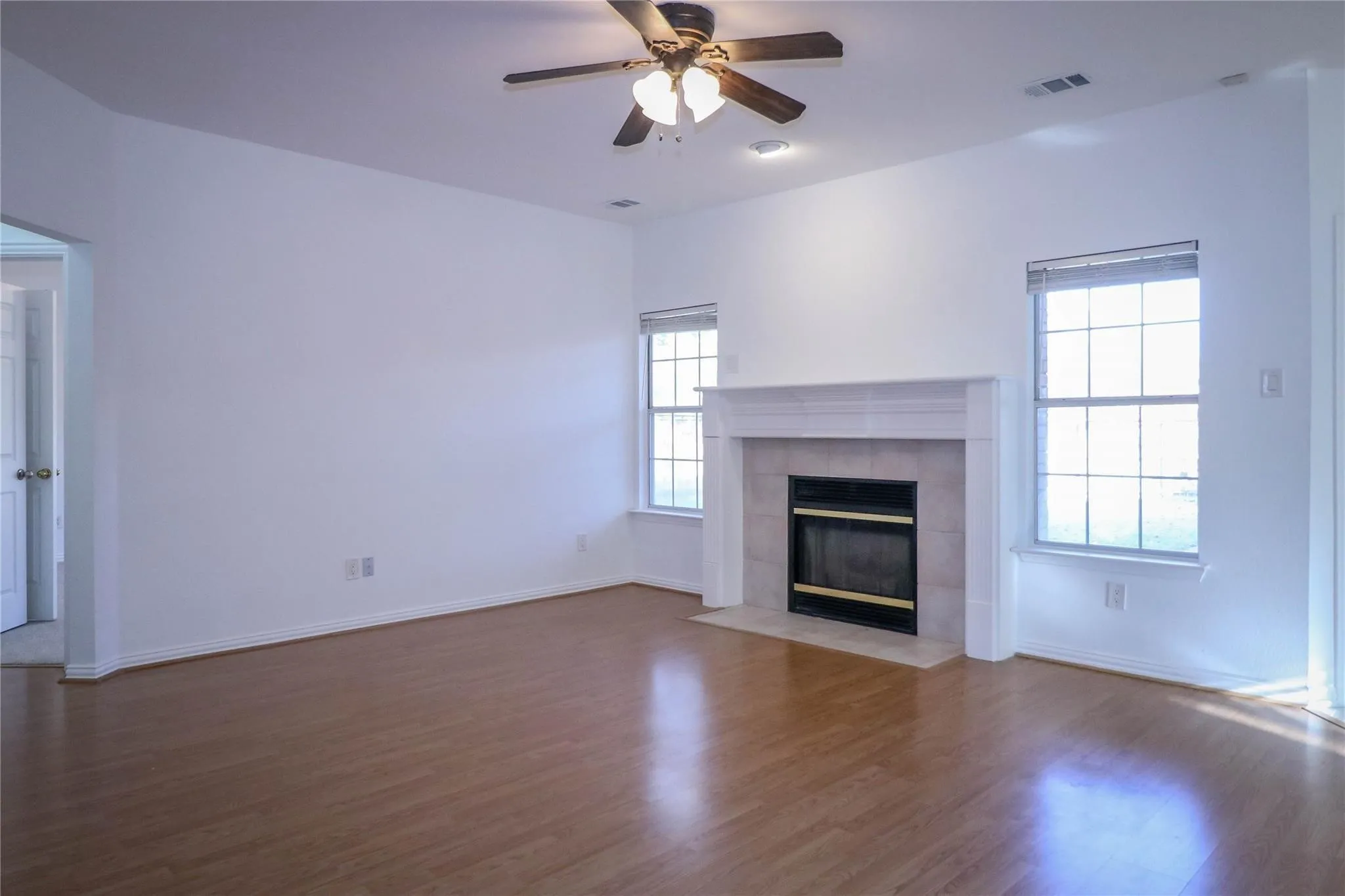 Living area featuring wood-finish flooring, a fireplace with a white mantel and light-toned surround, two windows, and a ceiling fan with integrated lighting