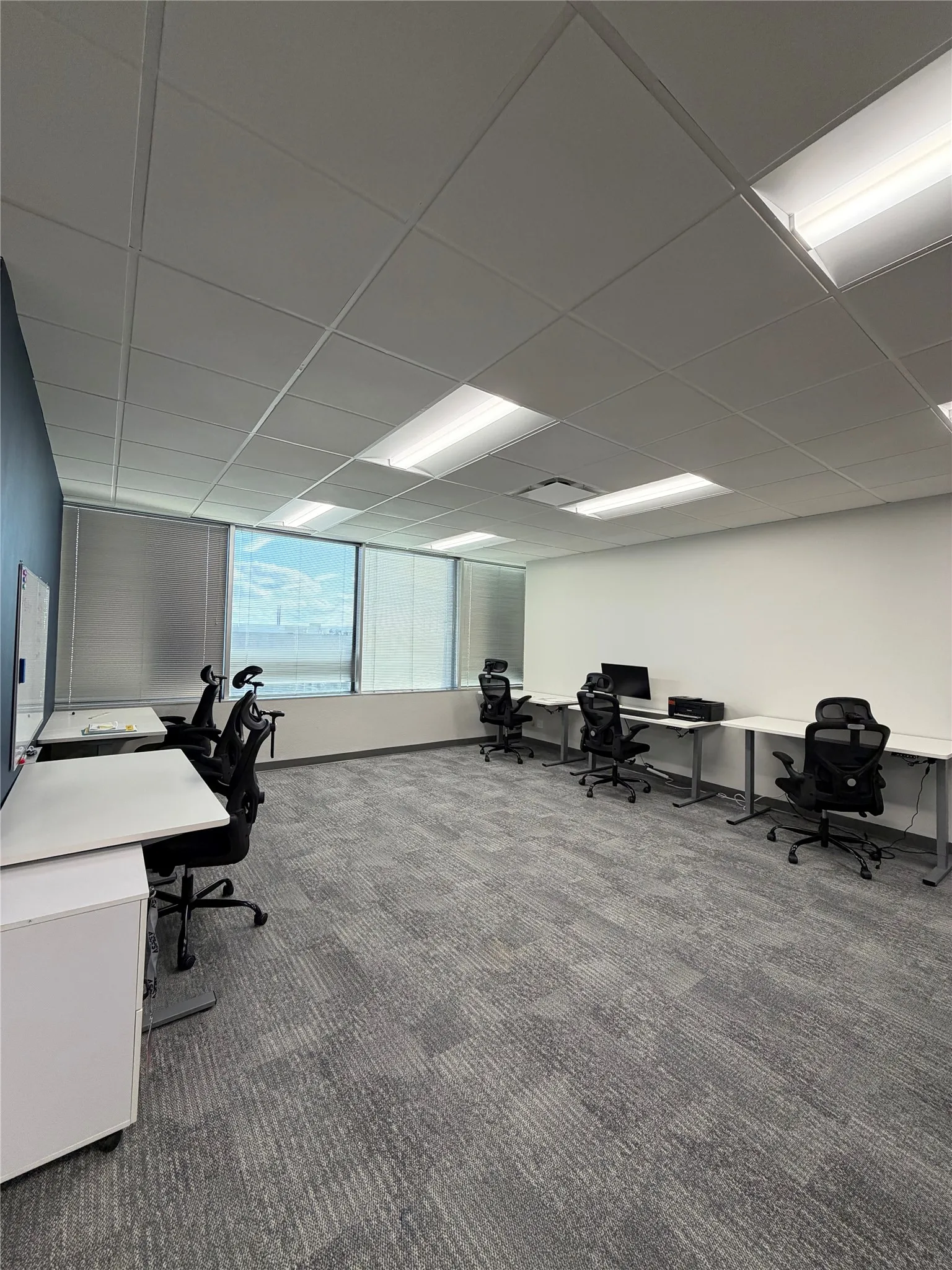 Large workspace featuring a suspended drop ceiling, recessed fluorescent lighting, wall-to-wall carpeting, and floor-to-ceiling windows with integrated blinds