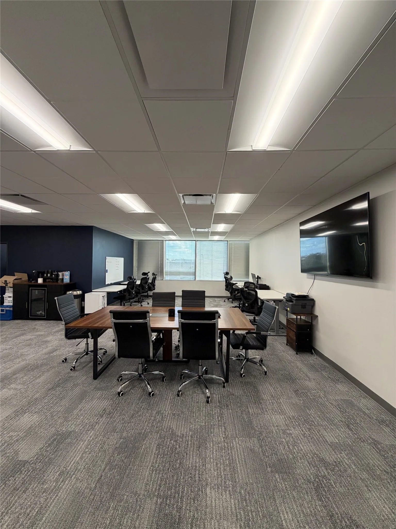 Interior space featuring a drop ceiling with recessed rectangular lighting, grey patterned carpeting, expansive window with natural light, and a built-in accent wall