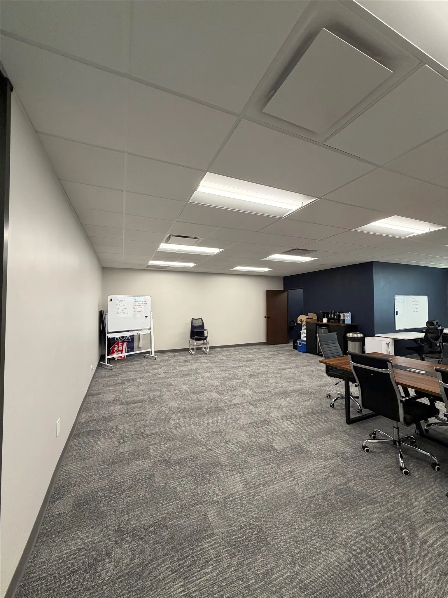 Open workspace featuring a drop ceiling with recessed fluorescent lighting, neutral-toned carpet tiles, and contrasting dark-toned accent walls