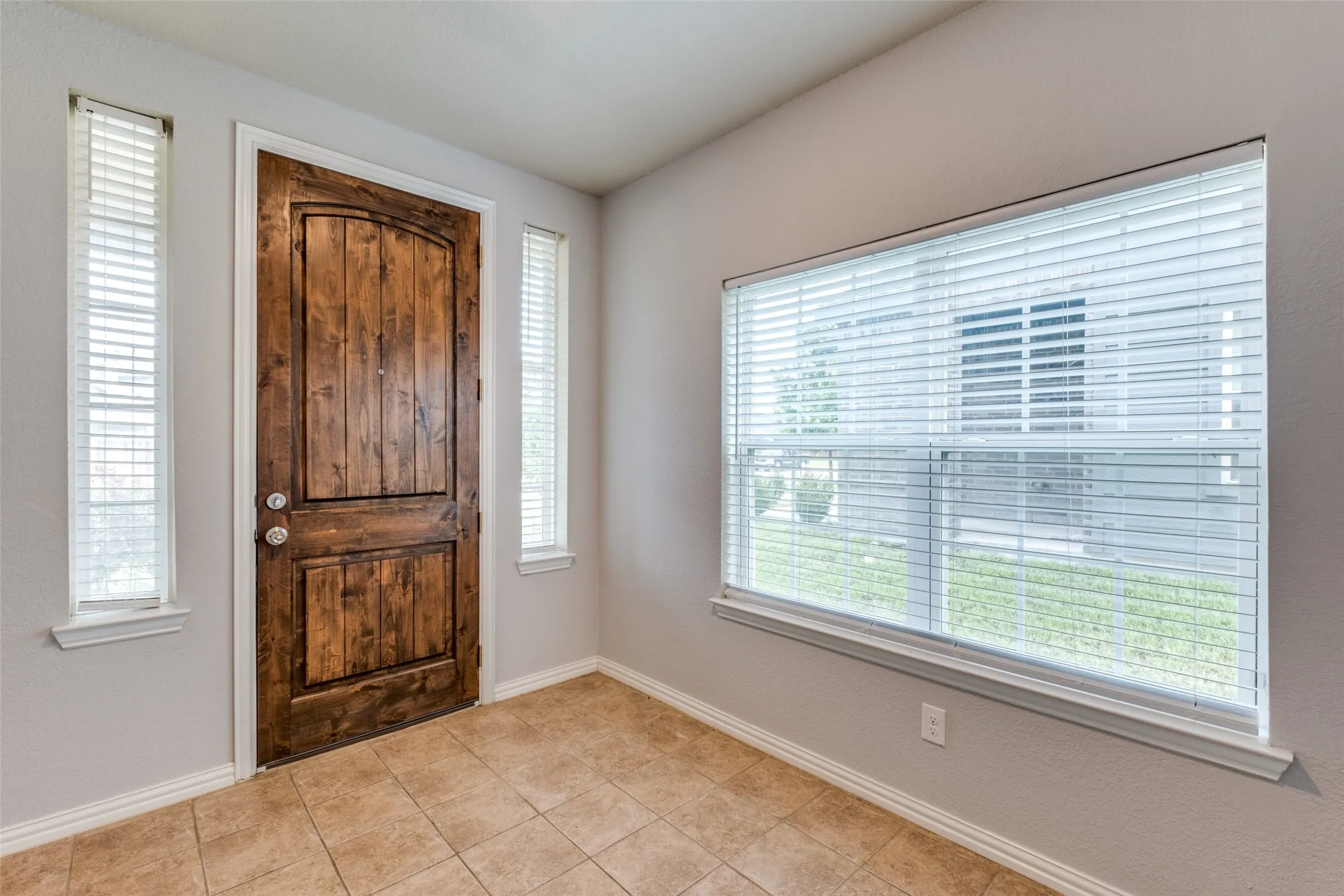 Entryway featuring a wood-finish arched-panel door, ceramic tile flooring, and multiple windows with horizontal blinds