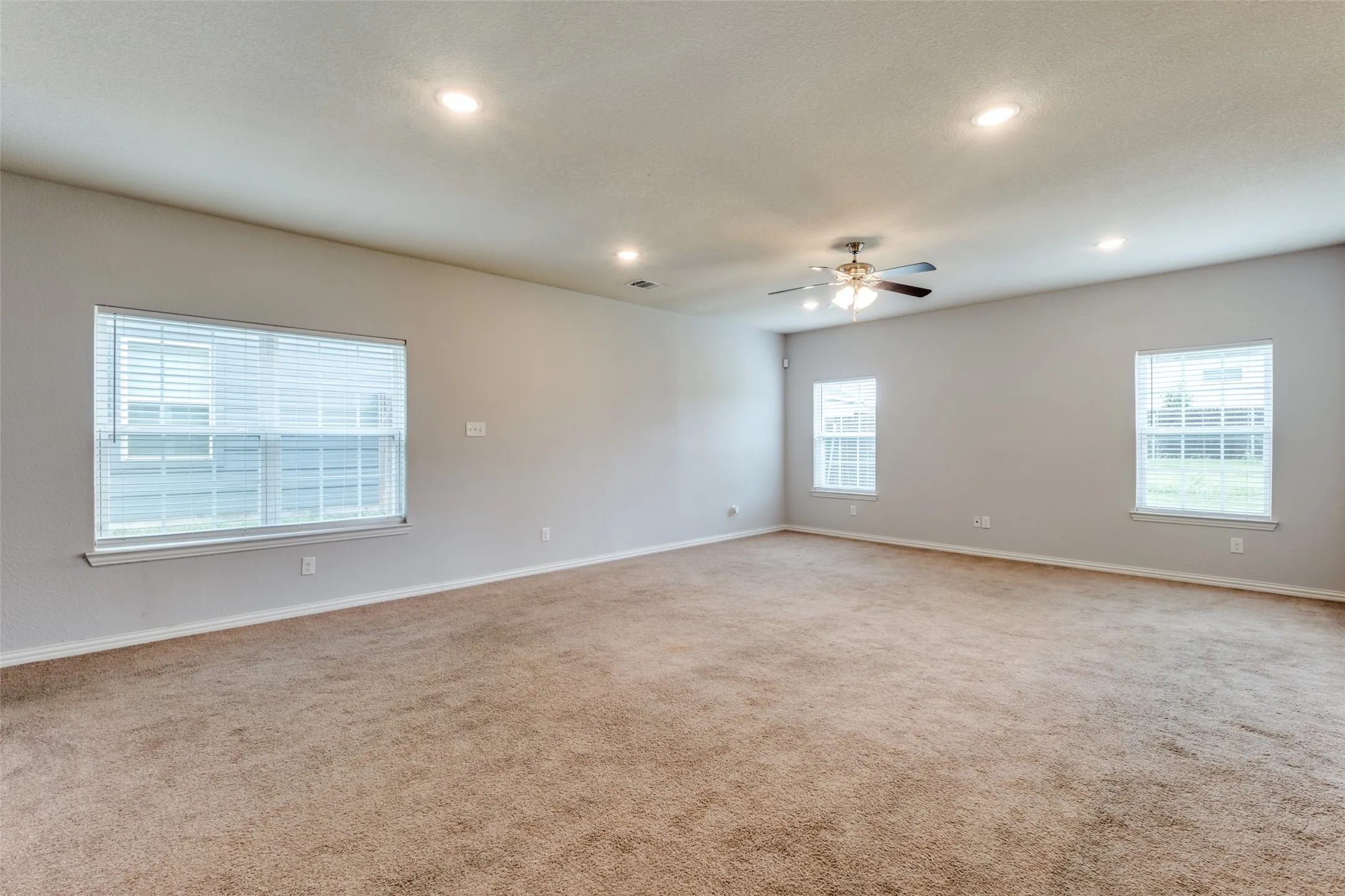 Spacious room featuring neutral carpeting, recessed lighting, a ceiling fan with integrated light fixture, and multiple windows with horizontal blinds