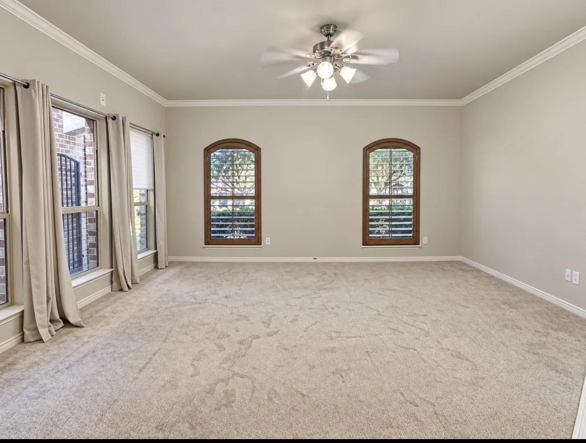 Neutral-toned interior featuring arched windows with wood-finish framing, crown molding, wall-to-wall carpeting, and a multi-light ceiling fan