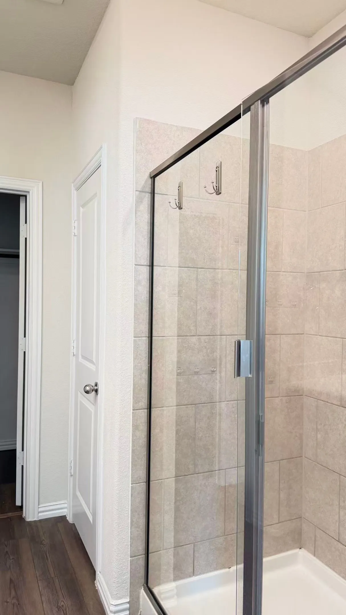 Shower enclosure featuring light-toned rectangular tile surround, semi-frameless glass door with brushed metal hardware, and wood-finish flooring