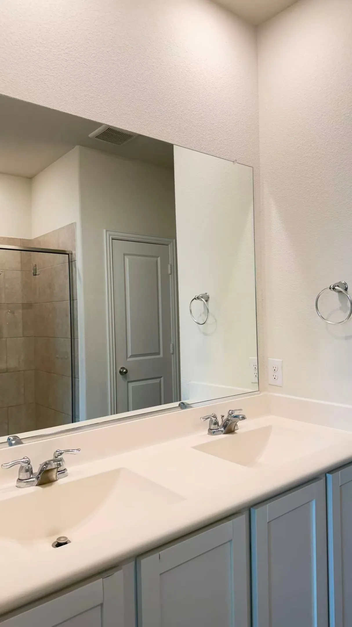 Double vanity featuring solid-surface countertops, integrated dual sinks, chrome dual-handle faucets, and gray Shaker-style cabinetry, complemented by a large wall-mounted mirror and two polished chrome ring towel holders