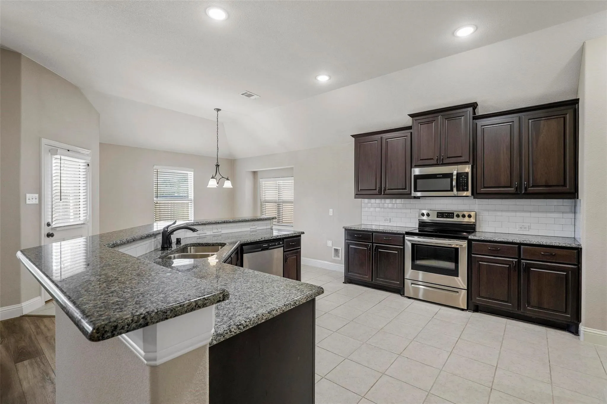 Kitchen featuring dark wood cabinetry, stone-finish countertops, stainless steel appliances, subway tile backsplash, and vaulted ceilings