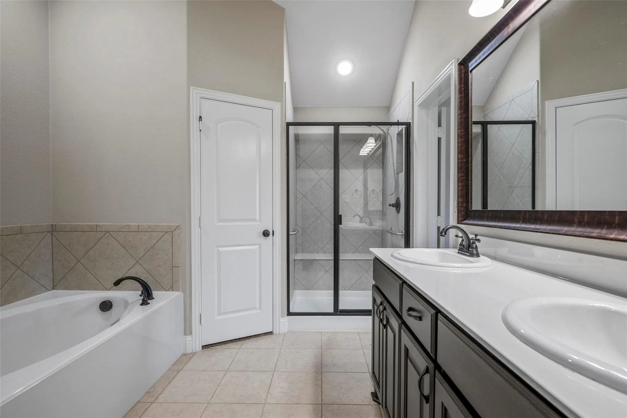 Dual-sink vanity with dark cabinetry and white countertops, corner soaking tub with tile surround, and a walk-in shower featuring a glass enclosure with a black frame