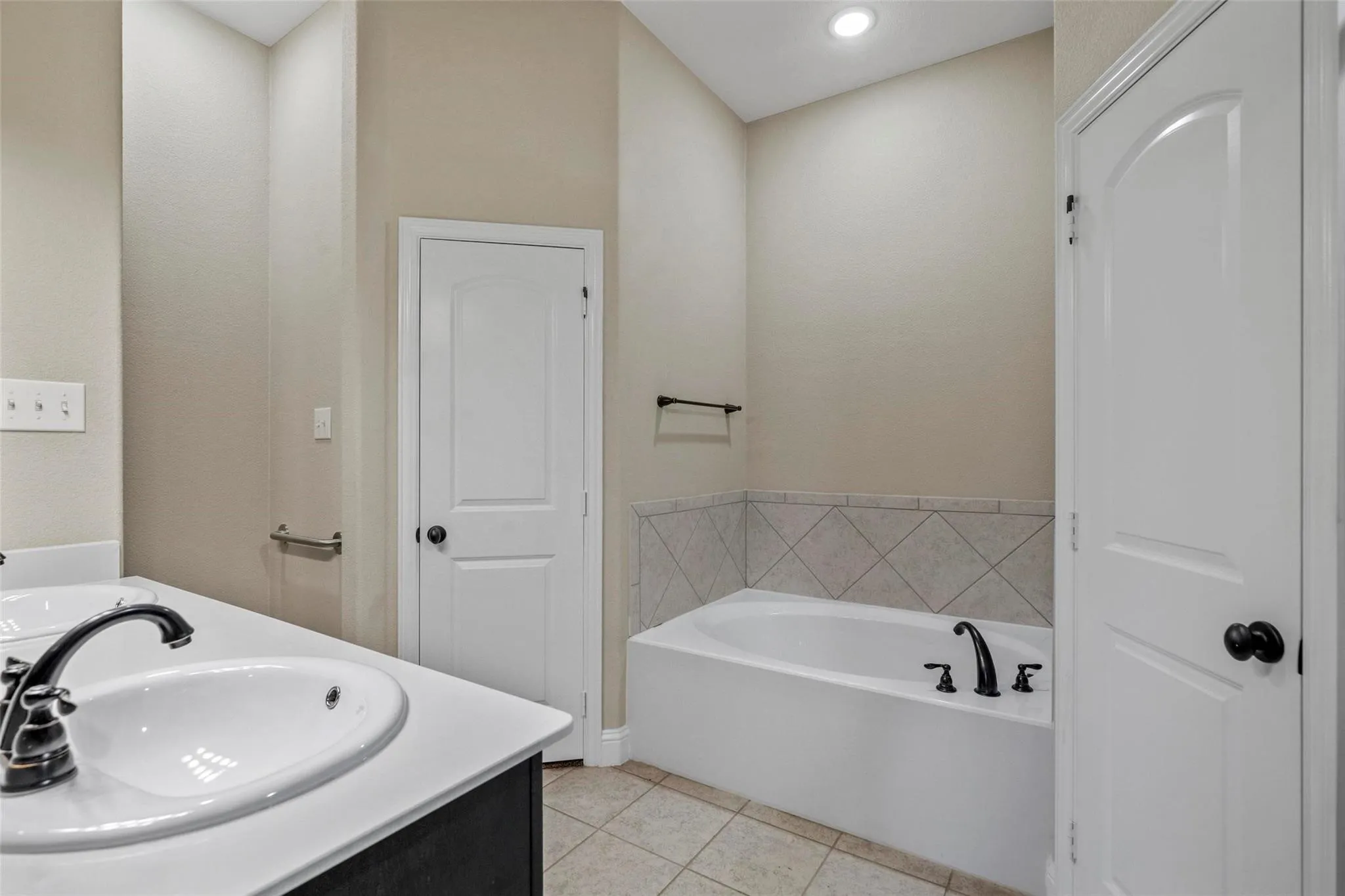 Deep-basin soaking tub with a tile surround, dual-sink vanity with a solid-surface countertop, recessed ceiling lighting, and ceramic tile​​‌​​​​‌​​‌‌​‌‌​​​‌‌​‌​‌​‌​​​‌​​ flooring