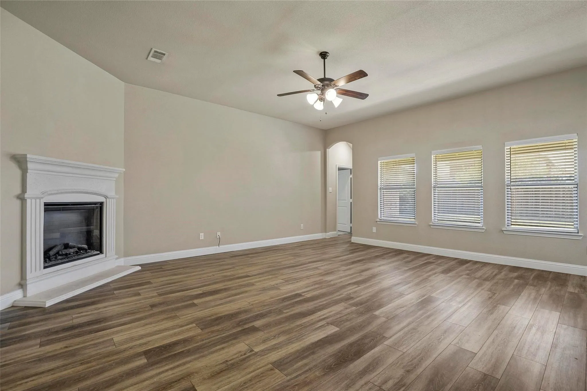 Open interior space featuring wood-finish flooring, a white mantel fireplace, three windows with blinds, and a ceiling fan with integrated lighting