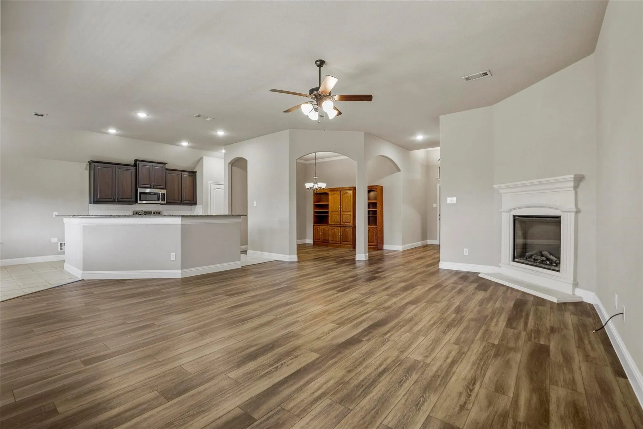Open-concept interior featuring wood-finish flooring, a corner fireplace with a white mantel, arched architectural entryways, a kitchen with dark cabinetry and a raised breakfast bar, and recessed lighting