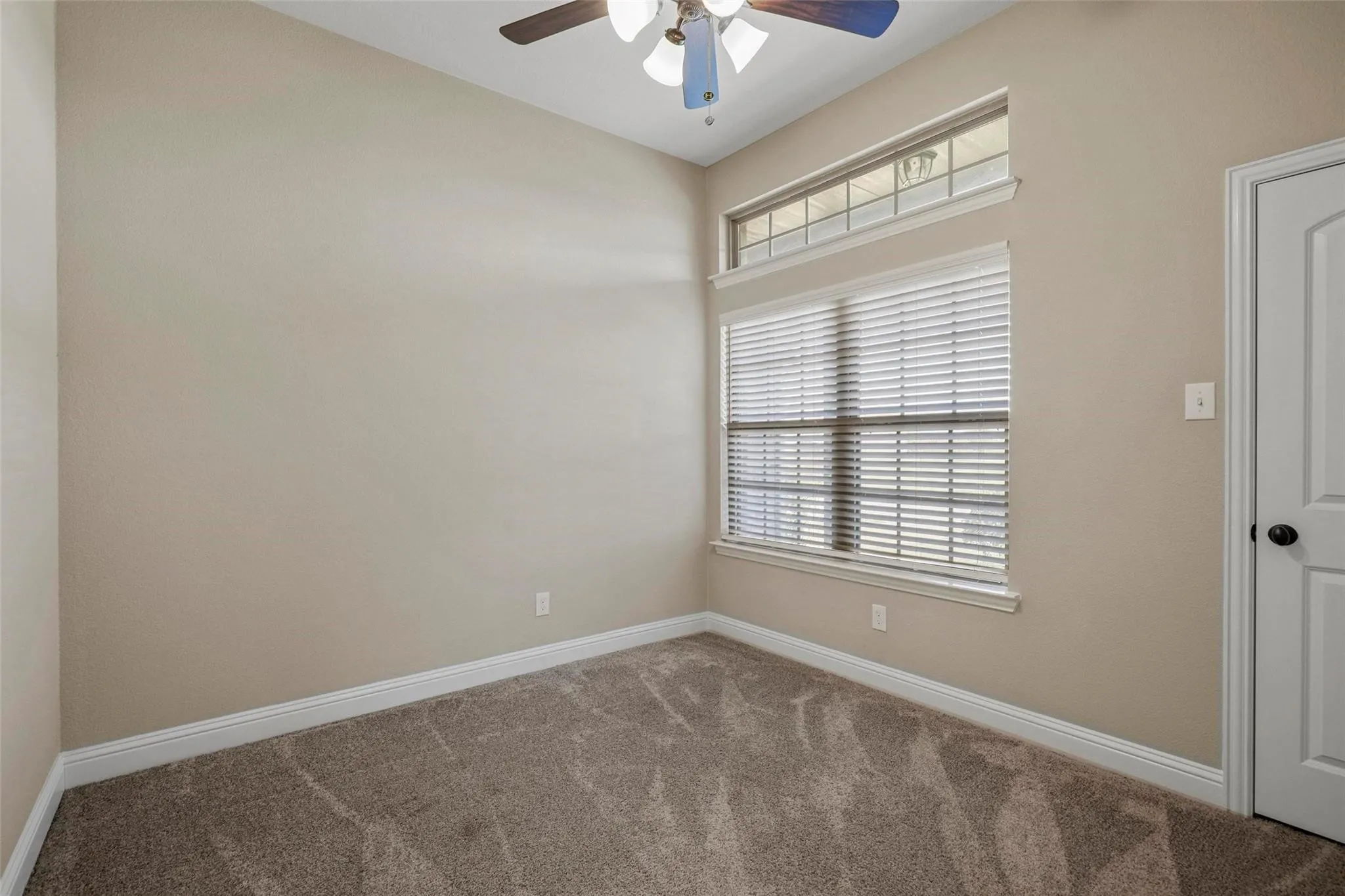 Neutral-toned interior room featuring carpeted flooring, a ceiling fan with integrated lighting, and a large window topped with a rectangular transom