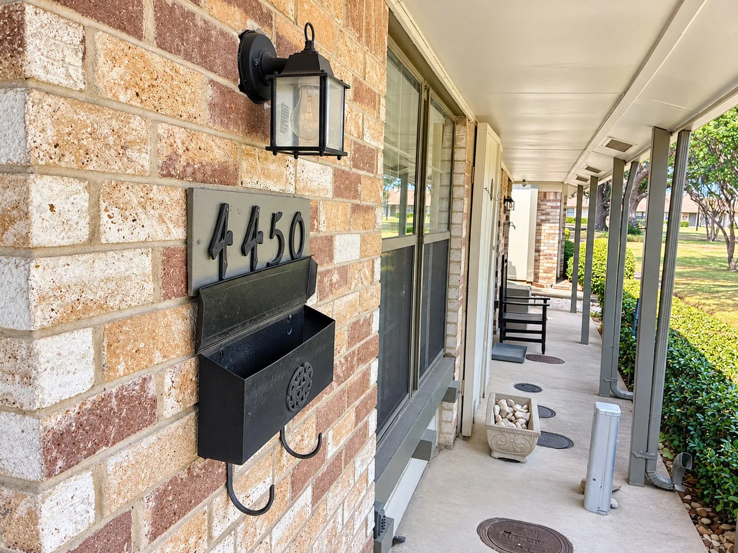 Covered front porch featuring a brick exterior facade, mounted metal mailbox with house numbering, and an exterior wall-mounted carriage light fixture