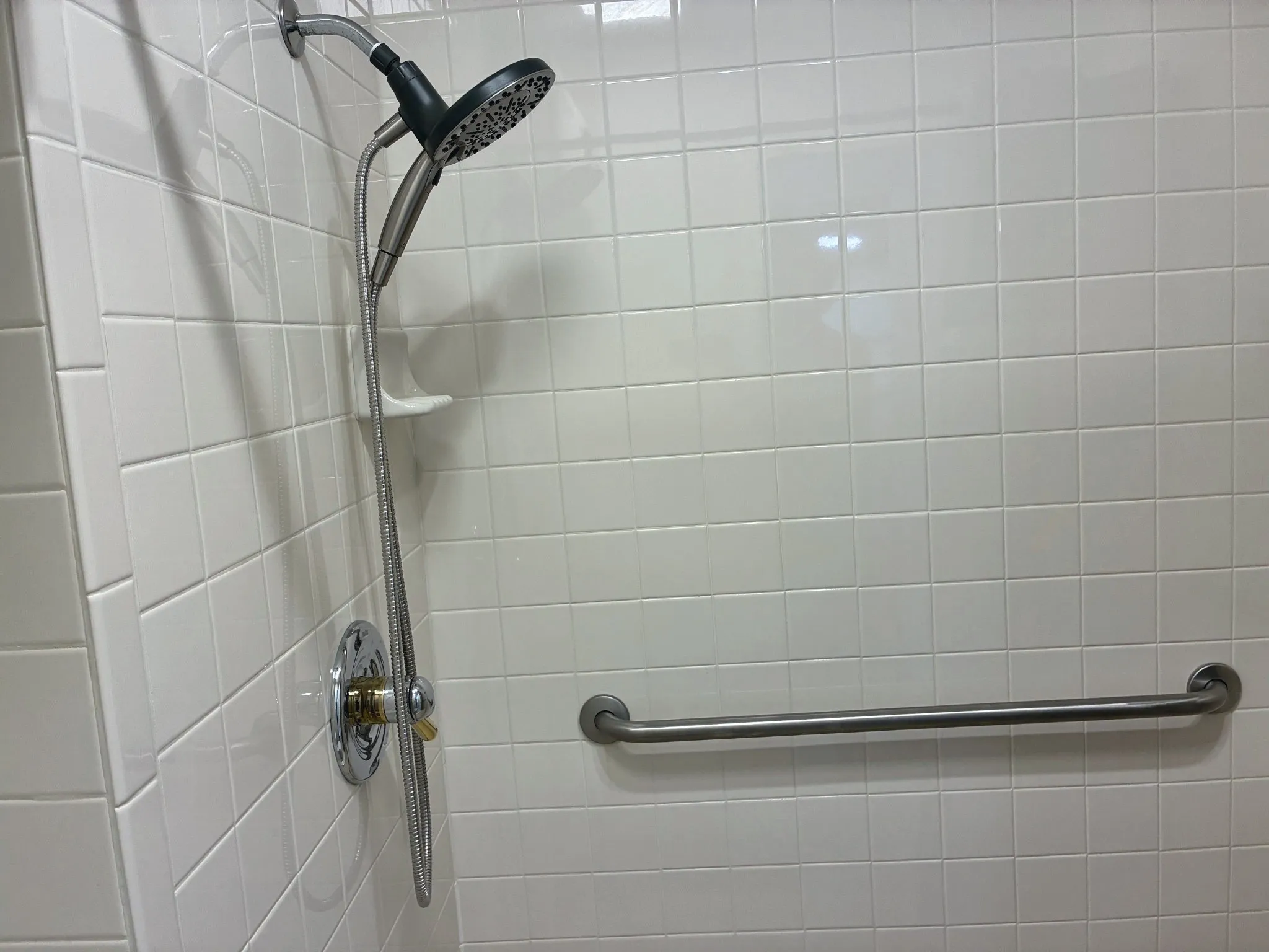 Shower enclosure featuring white square wall tile, a handheld showerhead, a built-in recessed soap dish, and a horizontal metal grab bar
