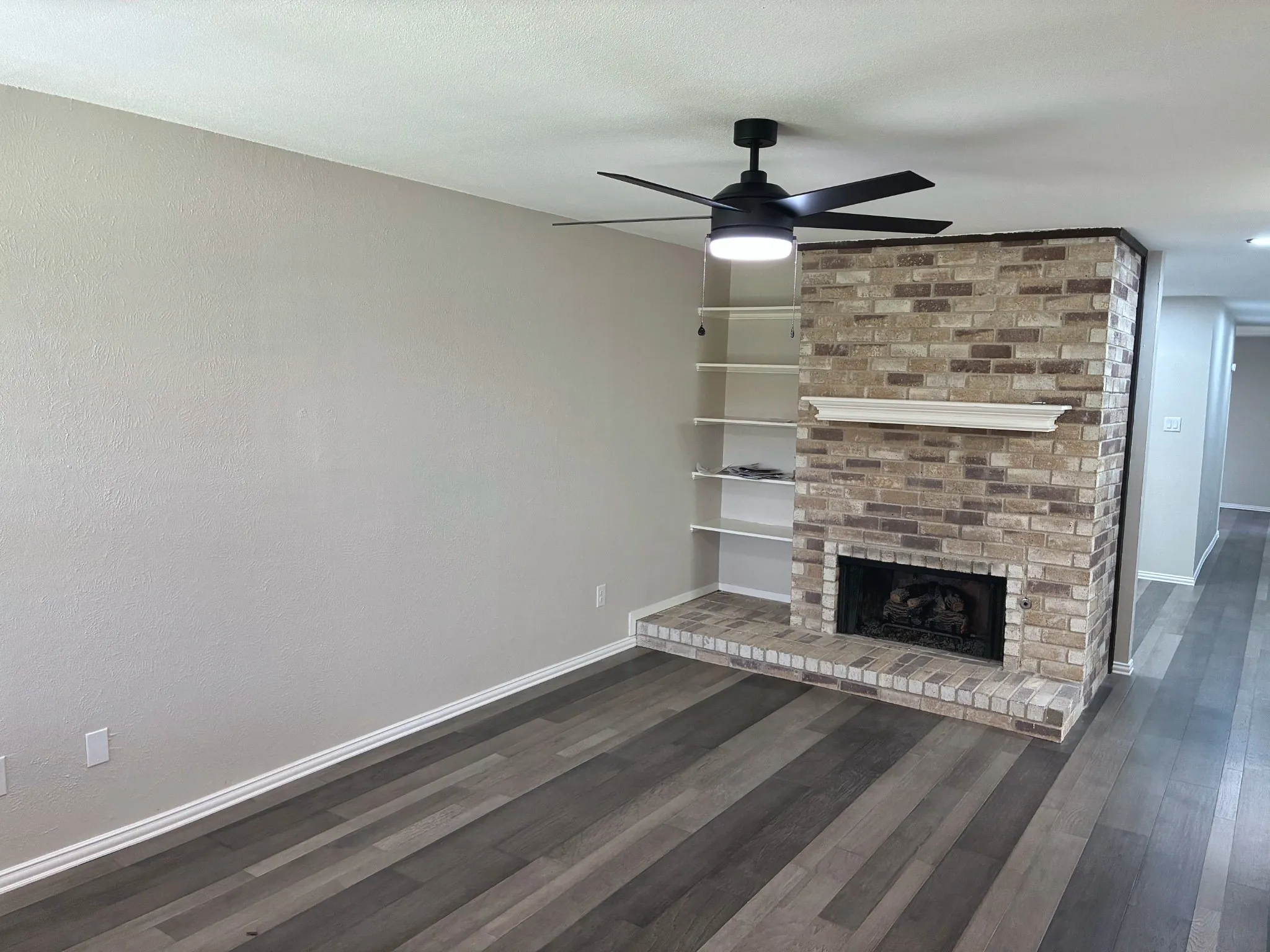 Brick fireplace with a mantel, integrated wall shelving, wood-finish flooring, and a ceiling-mounted light fixture with fan