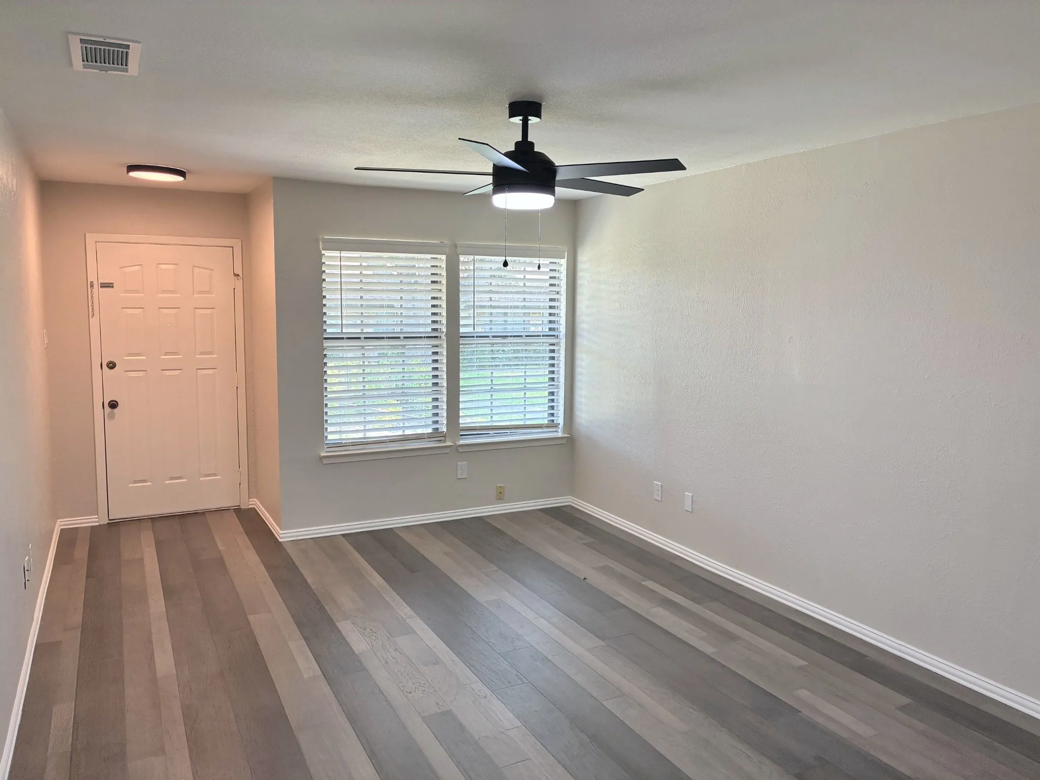 Room featuring wood-finish flooring, a matte black multi-blade ceiling fan with integrated lighting, dual pane windows with horizontal blinds, and a six-panel entry door
