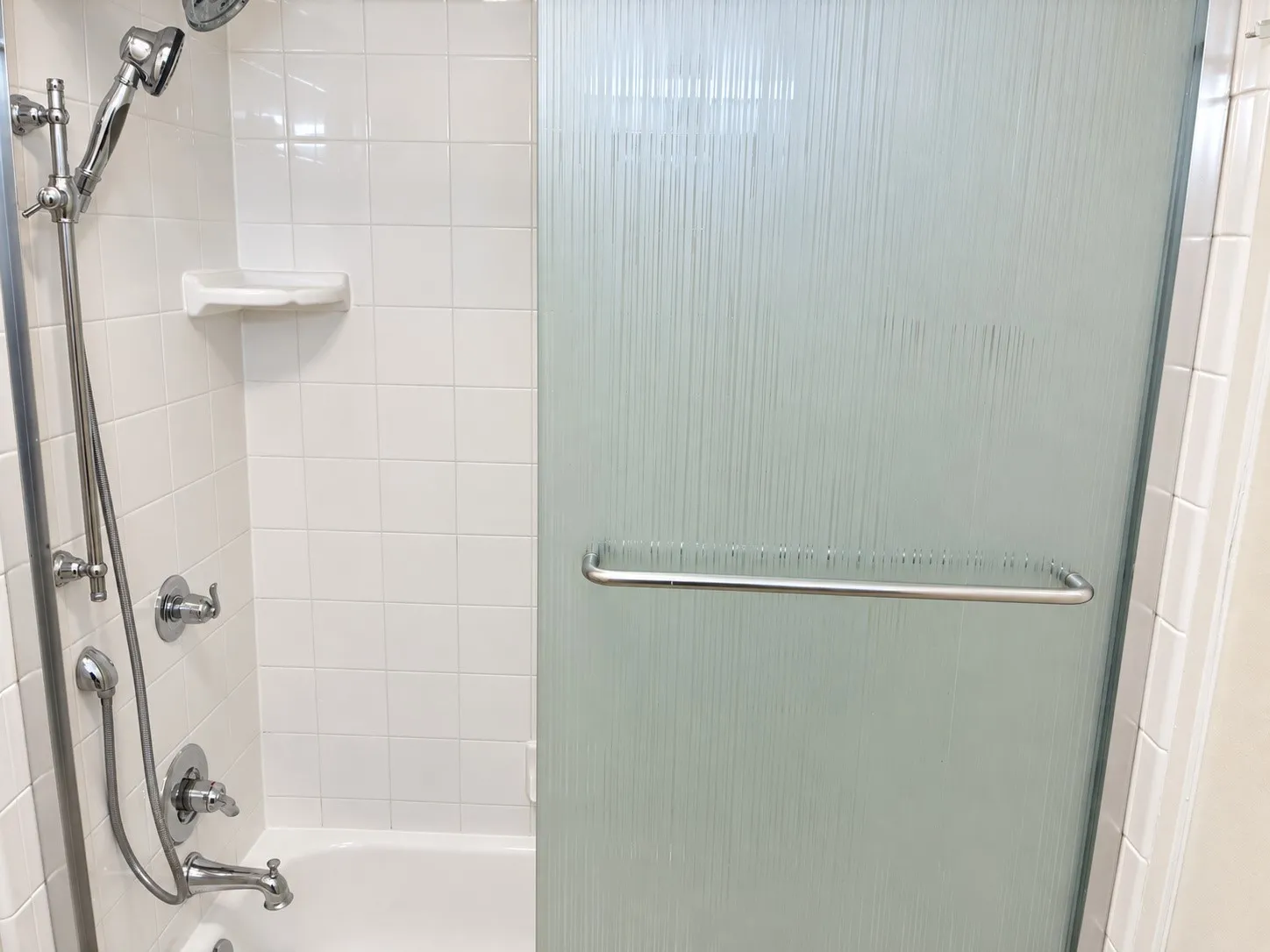 Shower enclosure featuring a chrome handheld shower head, integrated wall-mounted soap dish, textured glass sliding door with a horizontal chrome handle, and white tiled surround