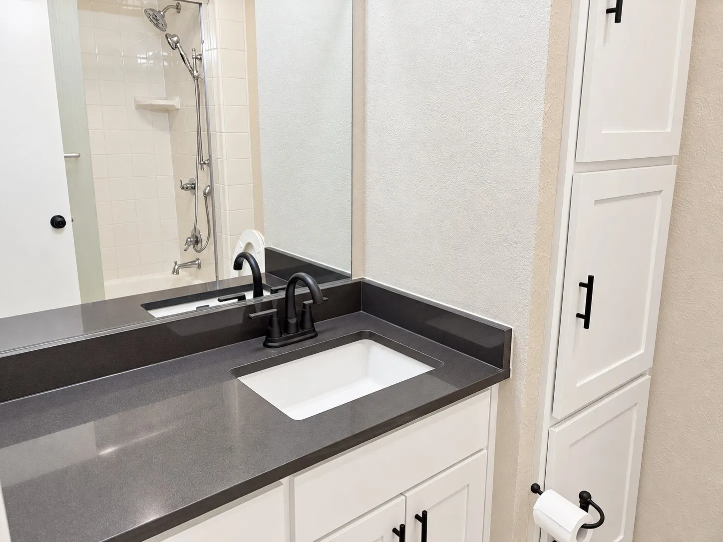 Bathroom vanity featuring a solid-surface countertop, an integrated rectangular sink, matte black faucet hardware, and white cabinetry with coordinating matte black pulls and built-in vertical storage