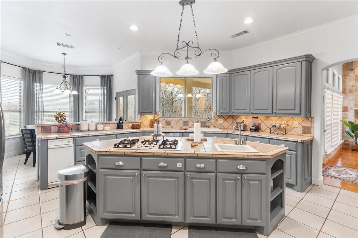 Kitchen featuring gray cabinetry, a central island with a built-in gas cooktop and sink, tile countertops with matching backsplash, and decorative pendant lighting