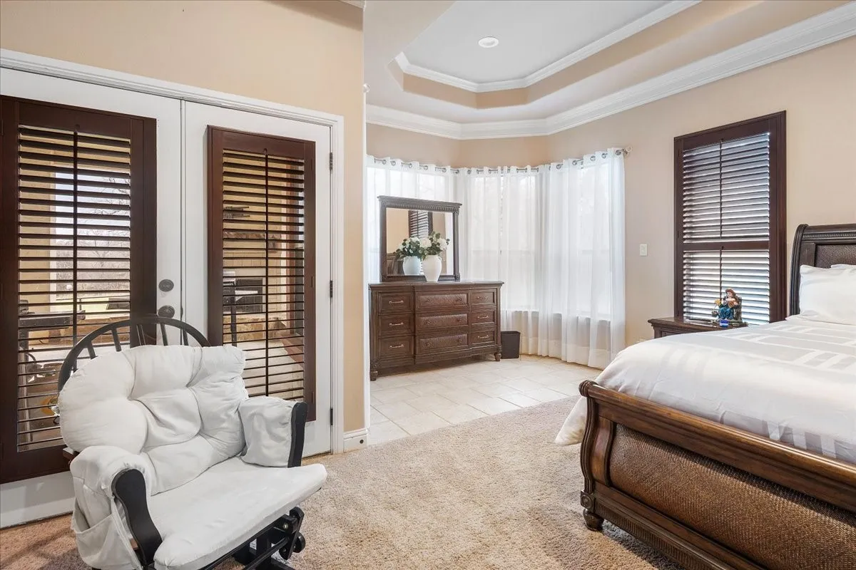 Tray ceiling, crown molding, double French doors with plantation shutters, multiple windows, and a combination of carpet and tile flooring