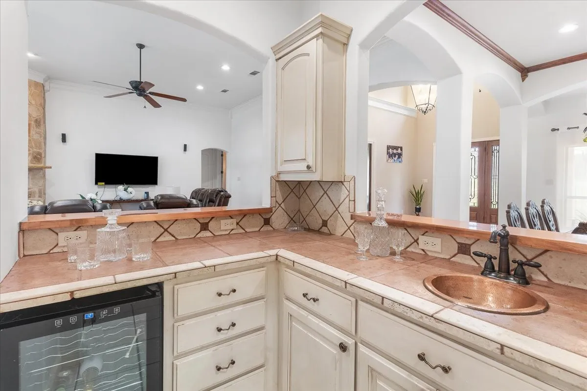 Kitchen wet bar featuring painted cabinetry, tile countertops with decorative accents, an integrated wine refrigerator, and a recessed copper basin with a metal faucet