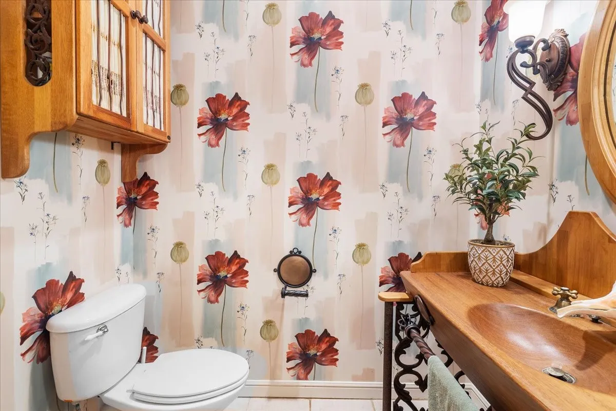 Powder room showcasing decorative wallpaper with floral and geometric patterning, white ceramic fixtures, wood-finish vanity with integrated sink, ornate mirror with wood frame, and decorative ceramic vessel accents