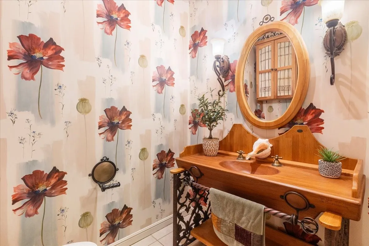 Charming guest powder bathroom featuring floral-patterned wallcovering, wood-finish vanity with integrated shelving, oval wood-framed mirror, and decorative wall-mounted fixtures