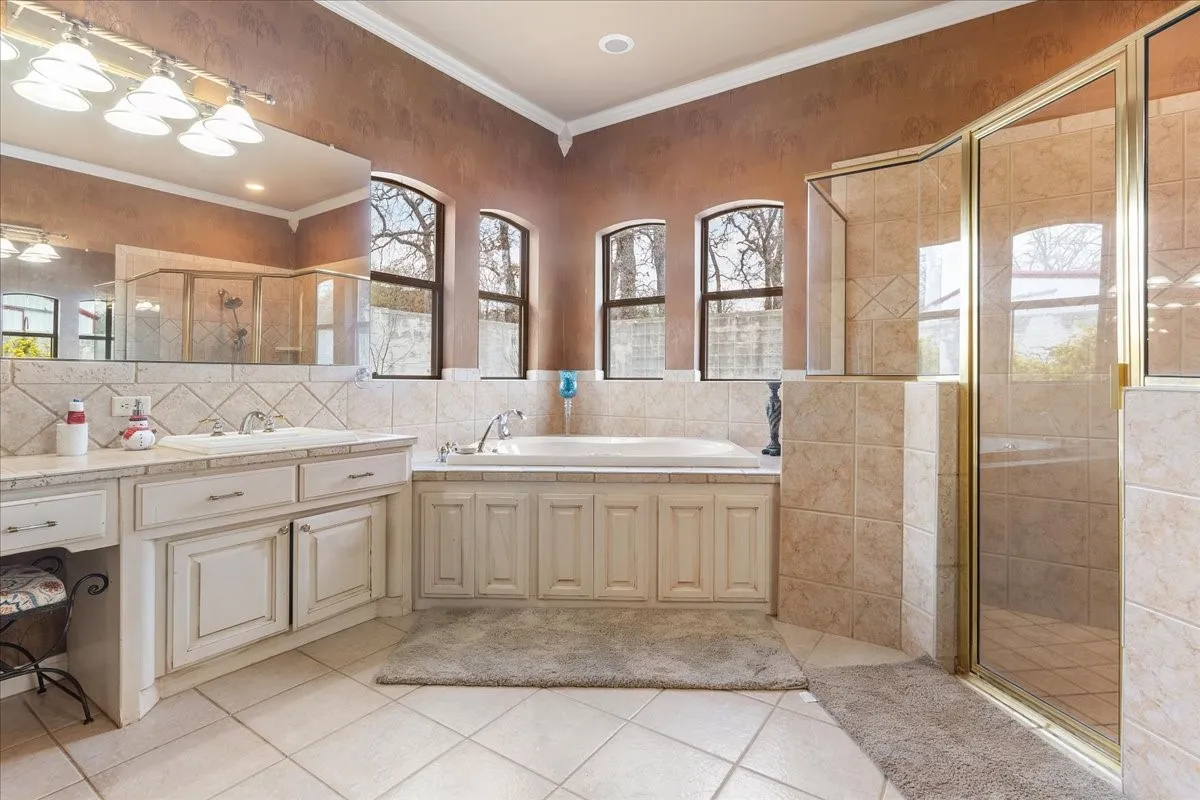 Bathroom featuring a dual-sink vanity with light cabinetry, tiled soaking tub surround, arched windows, and a glass-enclosed shower with tiled walls