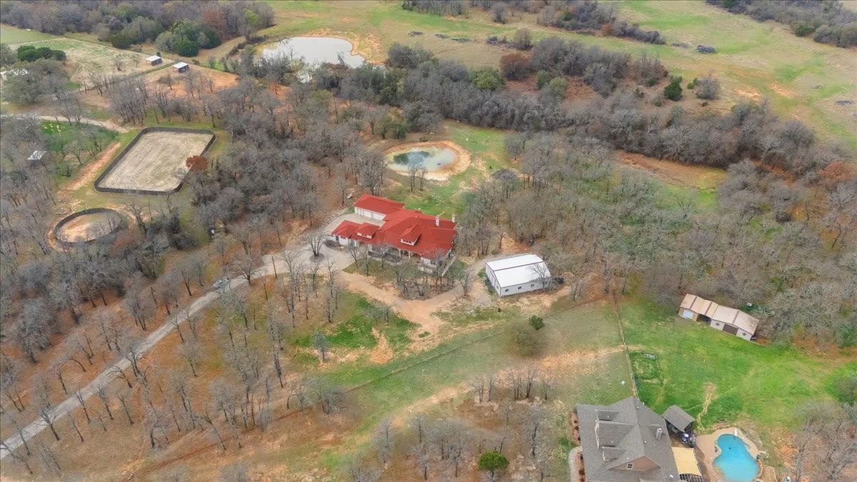 Aerial perspective of expansive acreage featuring a primary residence with a metal roof, dedicated equestrian arena, round pen, multiple outbuildings, and natural ponds