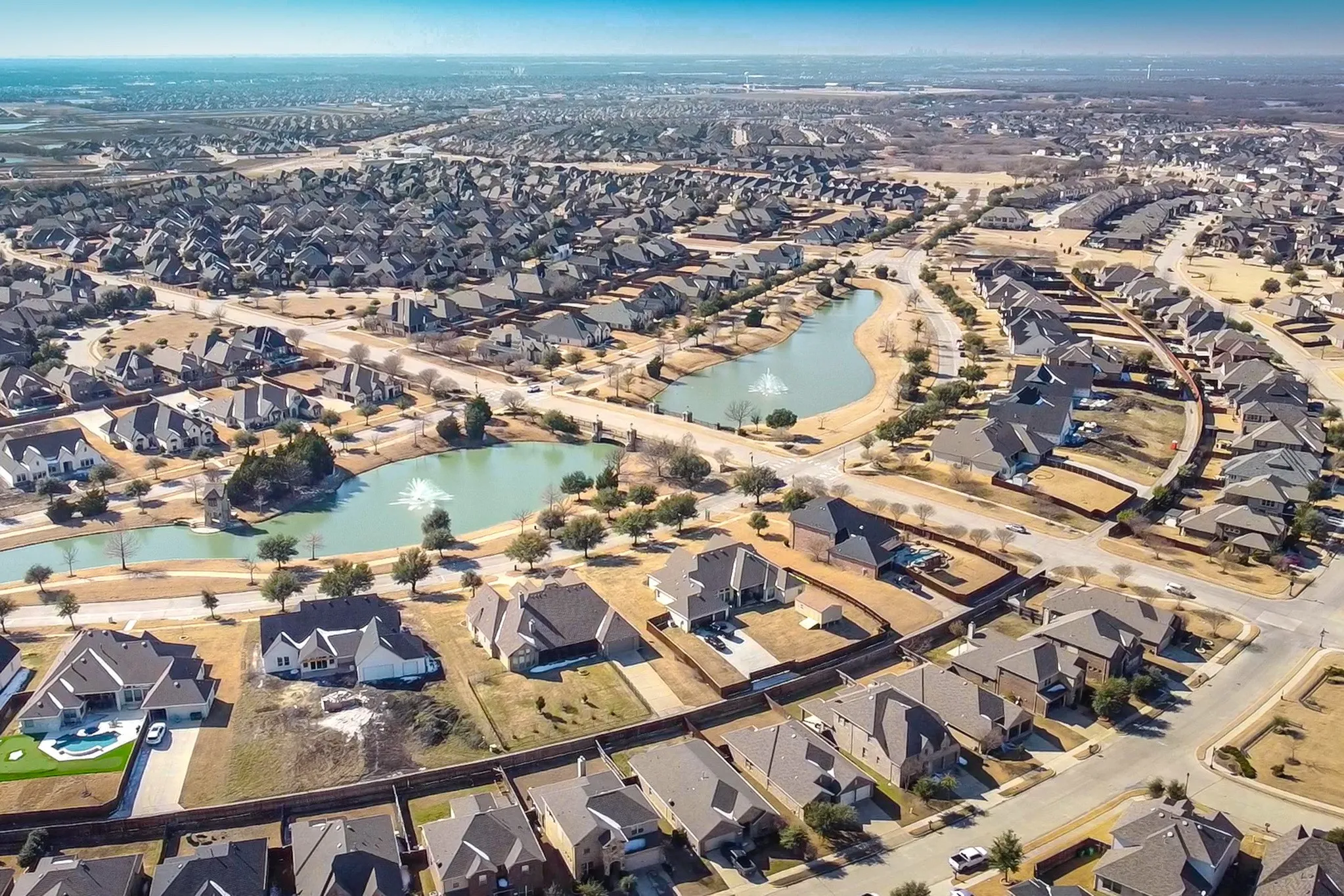 Aerial perspective of a suburban neighborhood featuring multiple retention ponds, decorative water fountains, paved street infrastructure, and residential structures with shingled roofing