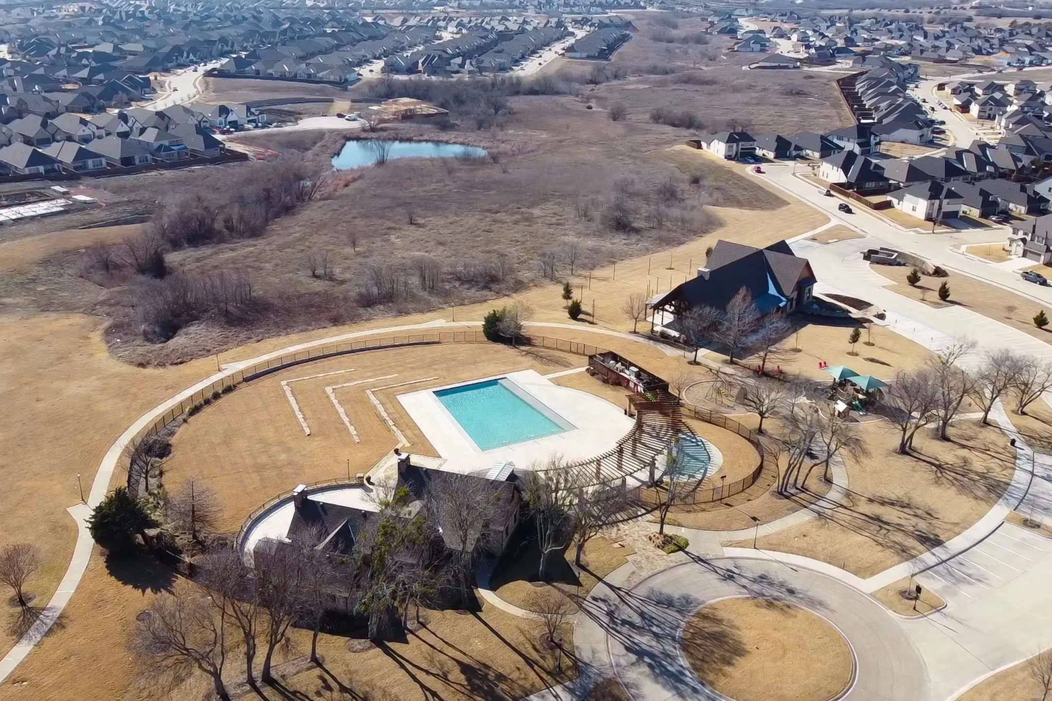 Aerial perspective featuring a rectangular swimming pool, expansive hardscaped patio, integrated wood-finish pergola, and fenced perimeter