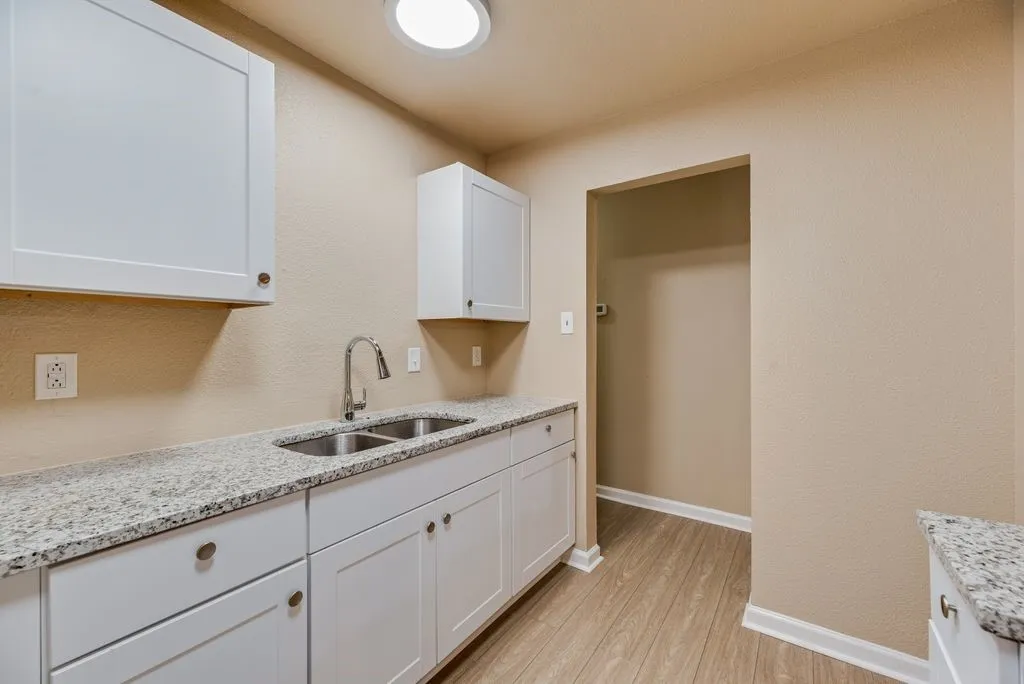 Utility area featuring white shaker-style cabinetry, stone-surface countertops, dual-basin stainless steel sink with gooseneck faucet, and wood-finish flooring
