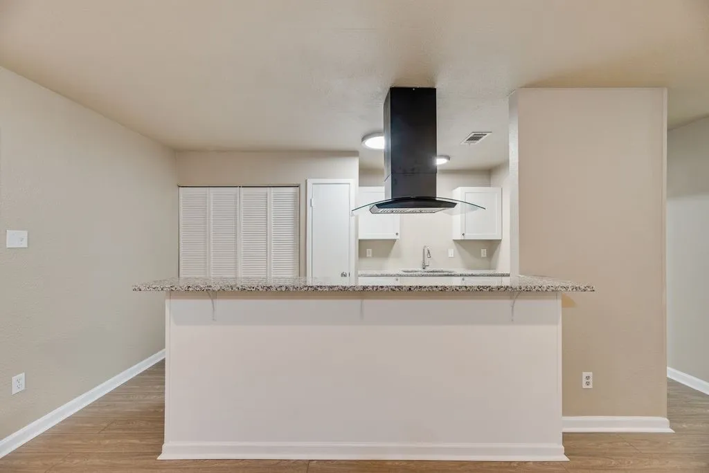 Kitchen area featuring a stone-pattern countertop breakfast bar, a black stainless steel chimney-style range hood, white shaker-style cabinetry, and wood-finish flooring