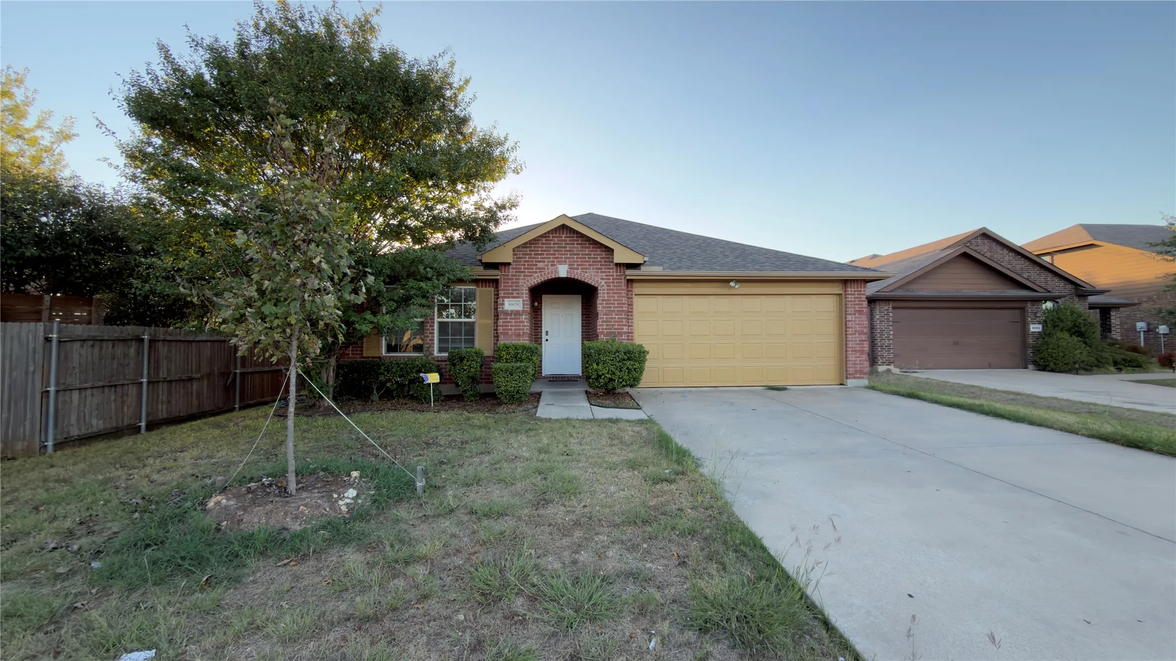 Single-story exterior featuring a brick facade, gabled roofline, concrete driveway, attached garage, and wood fencing