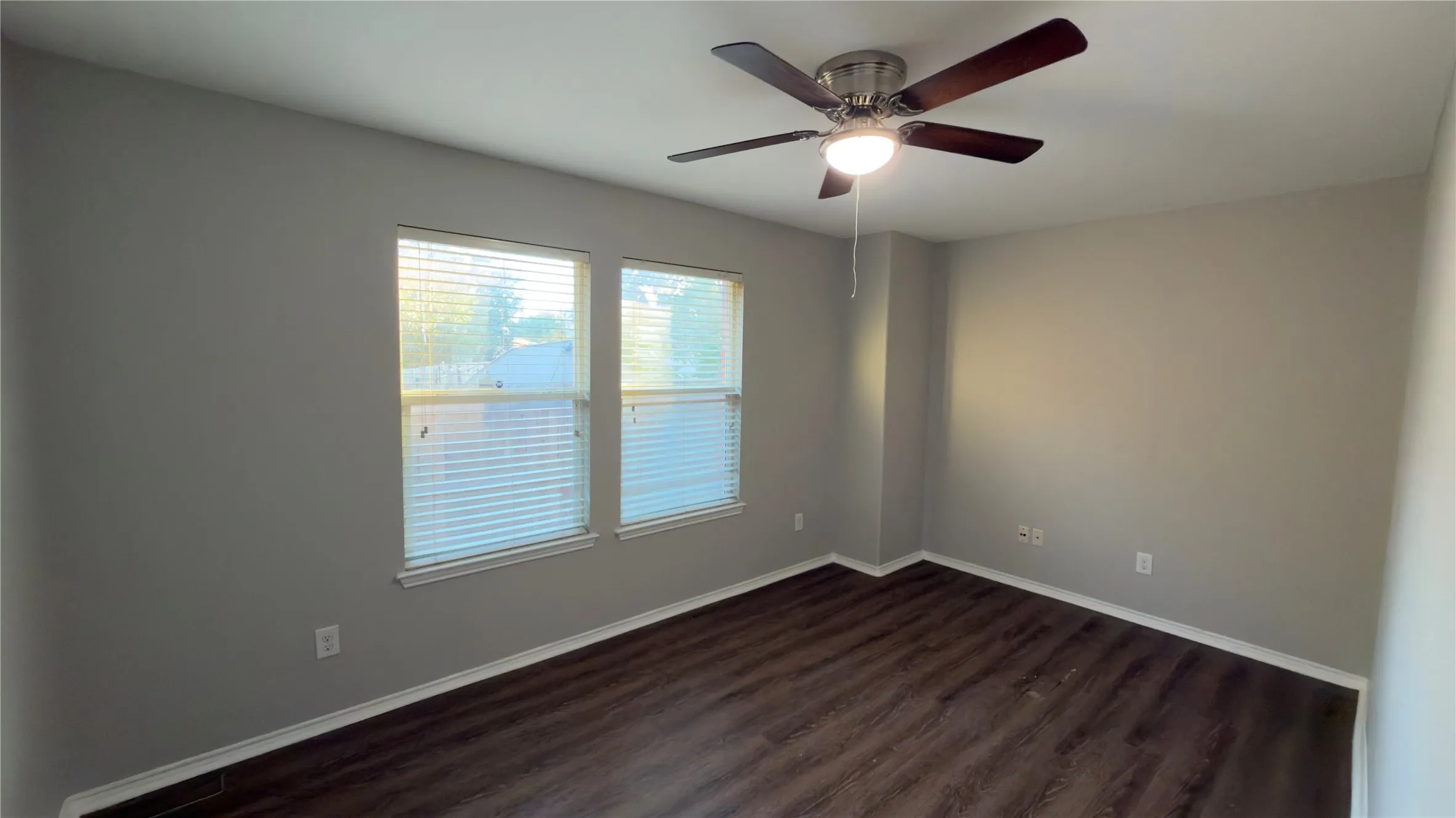 Neutral-toned interior featuring wood-finish flooring, dual windows with blinds, and a central ceiling fan with an integrated light fixture
