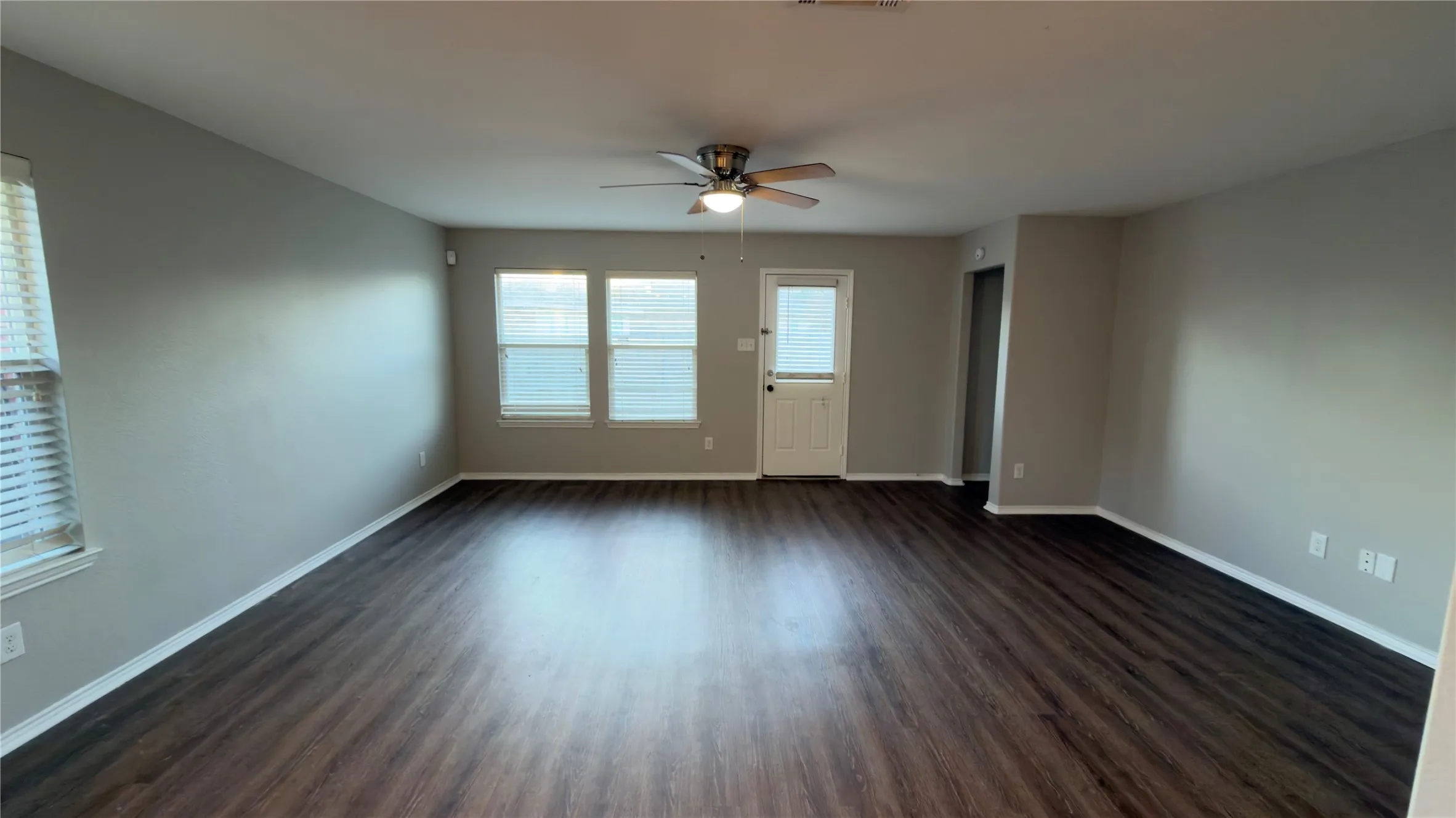 Open interior space featuring wood-finish flooring, neutral wall paint, a ceiling fan with integrated light fixture, and multiple windows with horizontal blinds