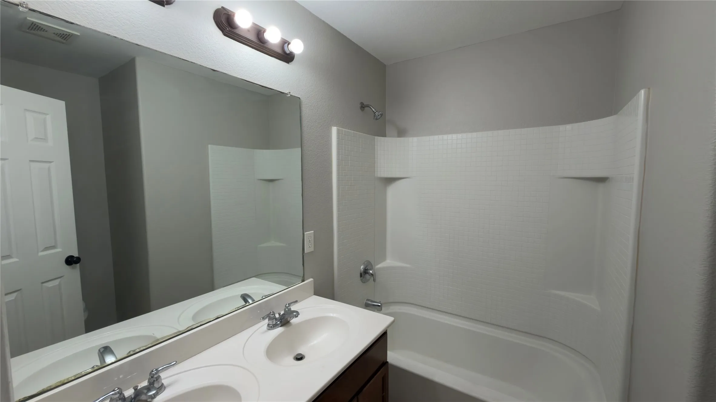 Double-sink vanity with a solid-surface countertop, full-width vanity mirror, triple-bulb light fixture, and a tub-shower combination featuring a molded surround