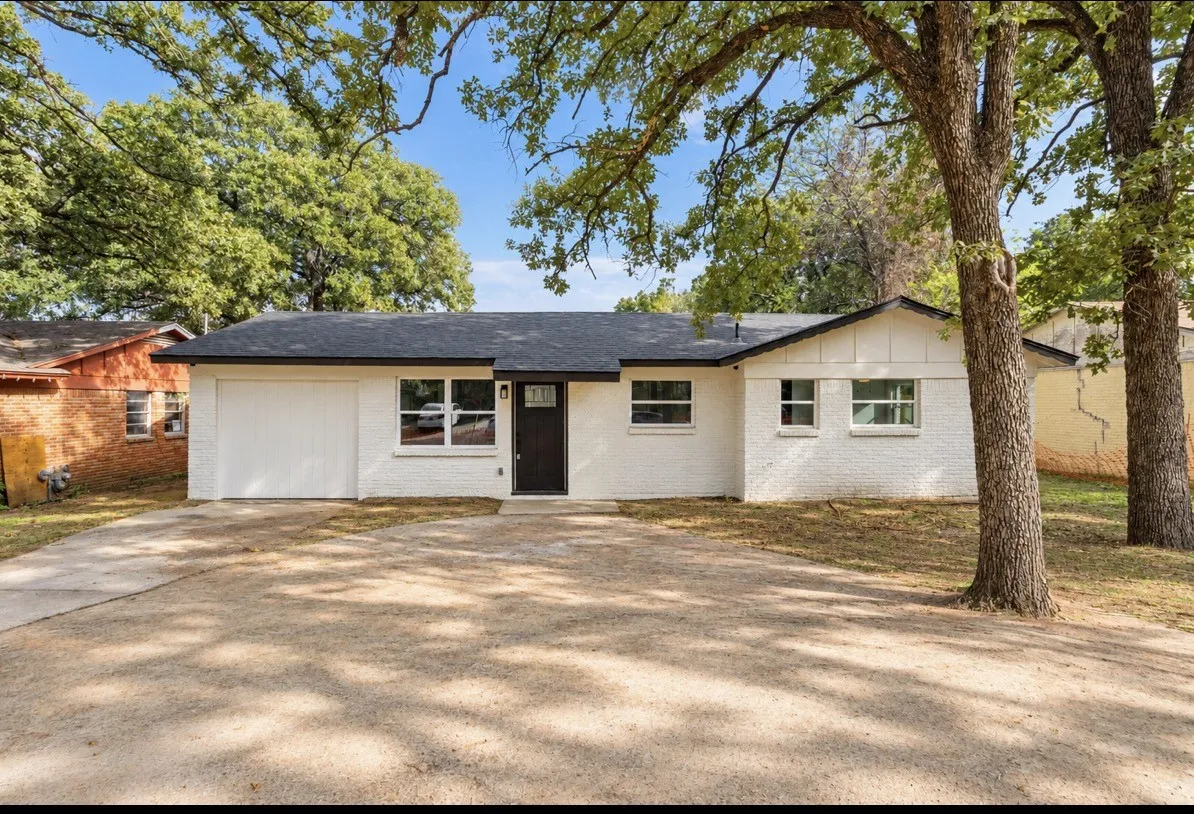 Single-story residence featuring a painted brick exterior, a composition shingle roof, an attached garage, and mature hardwood trees