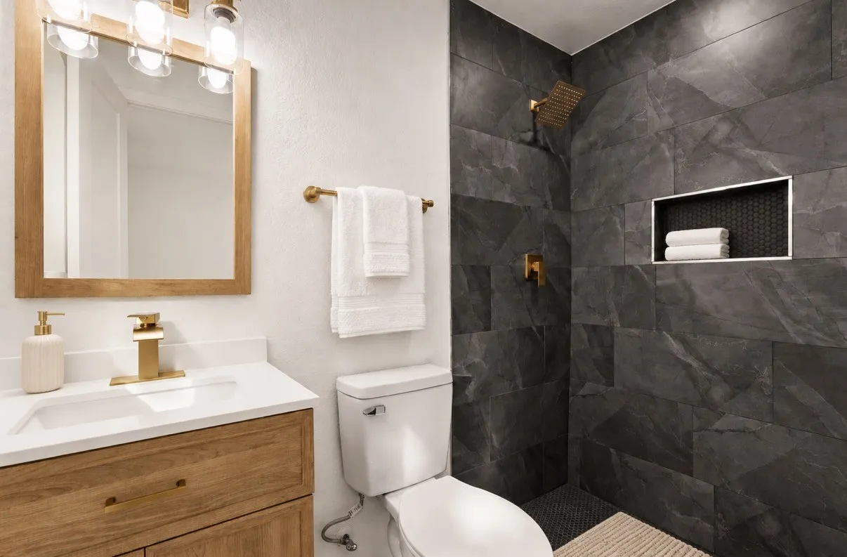 Wood-finish vanity with a stone-top integrated sink, brass-tone hardware, large-format dark stone shower tile, and built-in shower niche