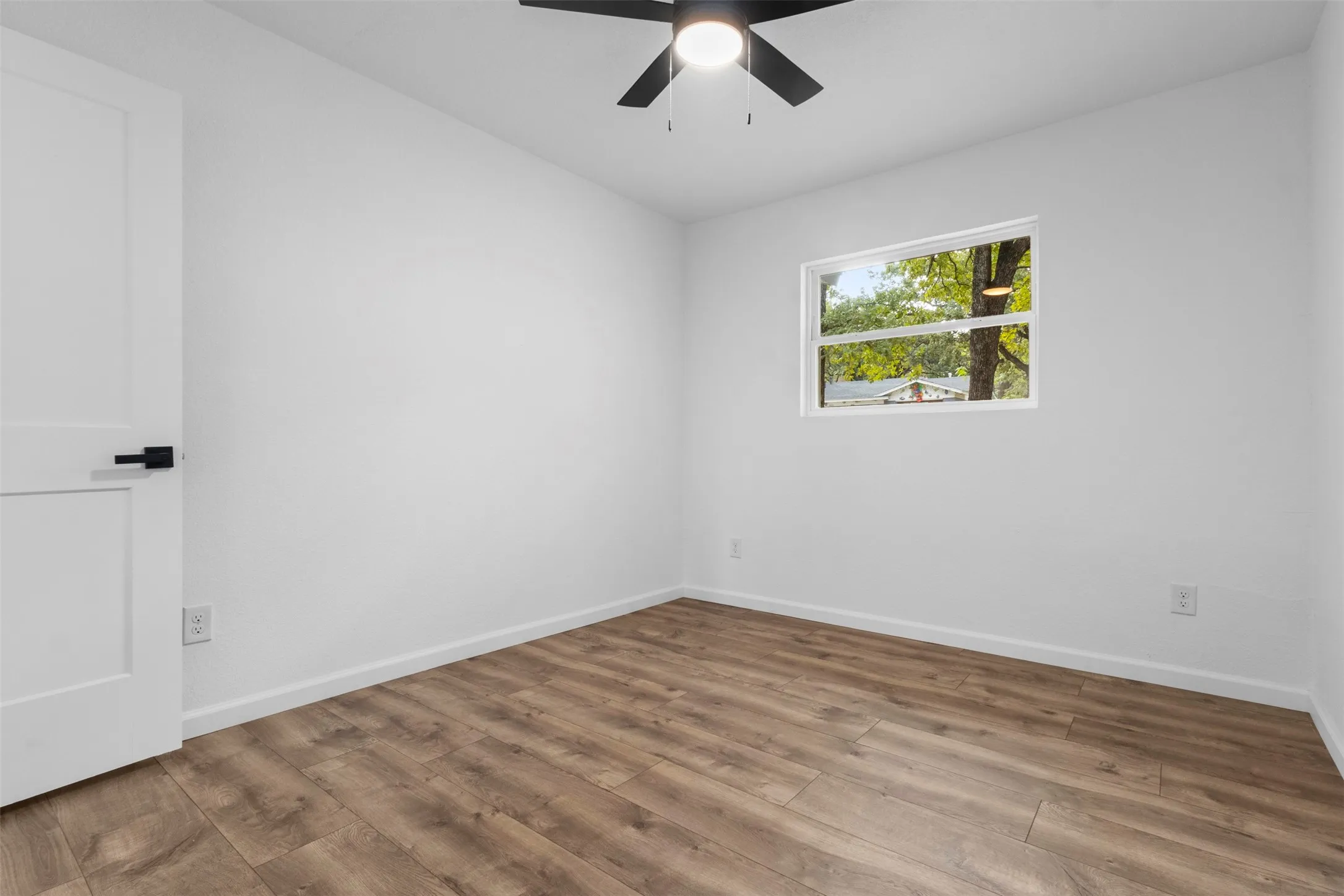 Room featuring wood-finish flooring, a matte black ceiling fan with an integrated light fixture, a single-panel interior door with matte black hardware, and a single-pane window