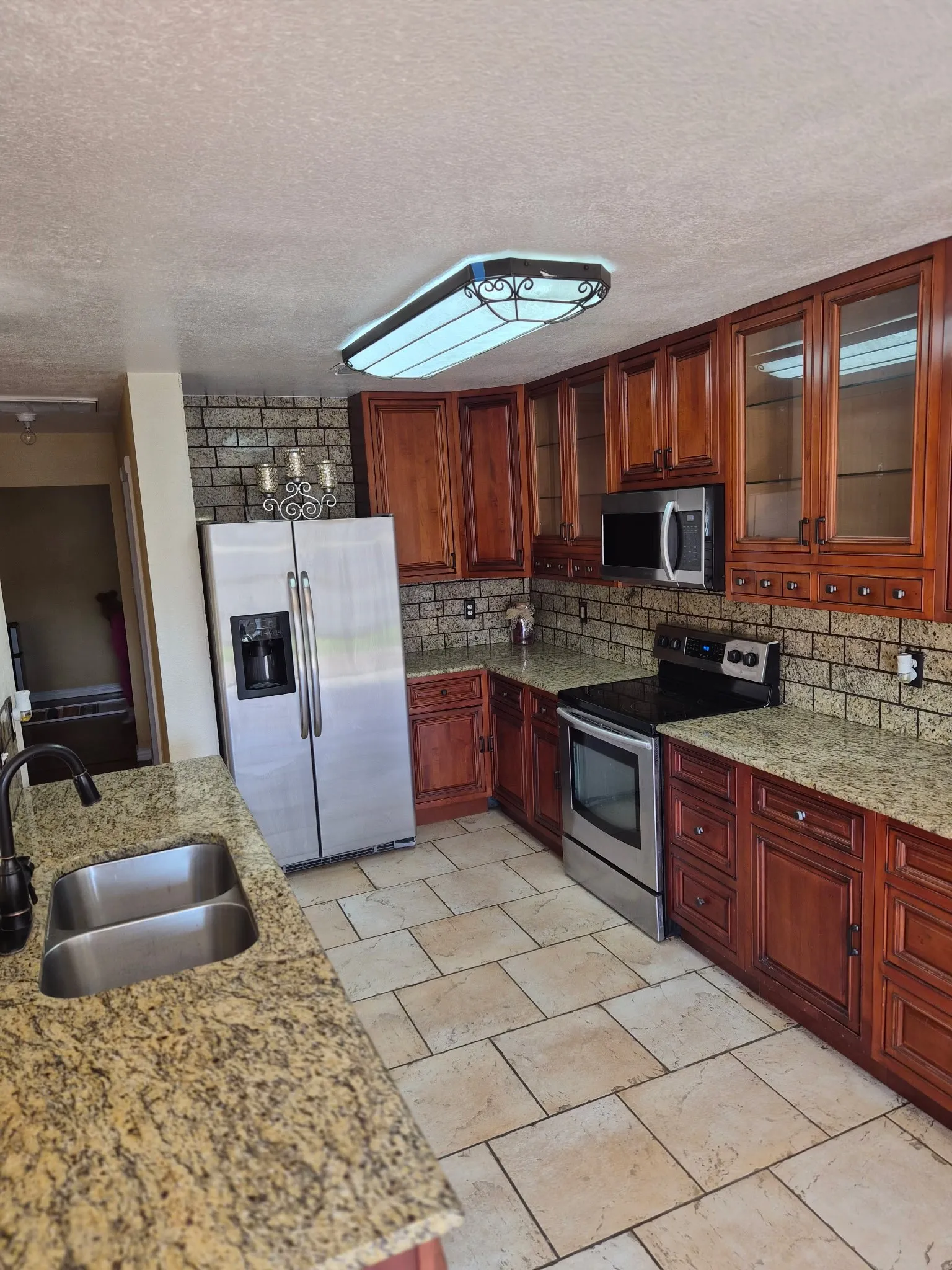 Kitchen featuring wood cabinetry with glass-panel upper doors, stone-finish countertops, tile backsplash, stainless steel appliances, and tile flooring