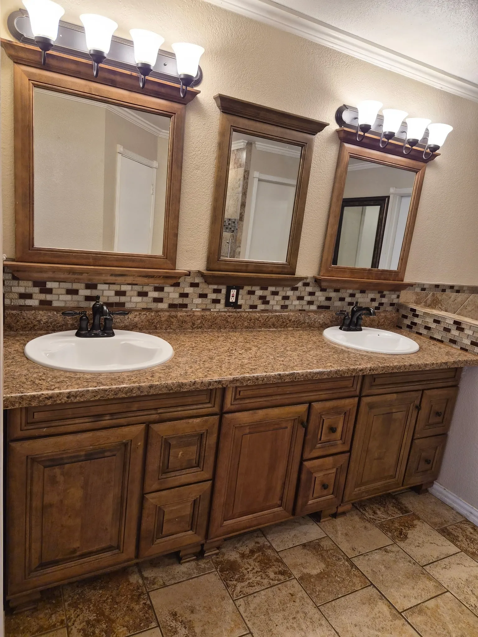 Dual-sink vanity featuring stained wood cabinetry, stone-finish countertops, mosaic tile backsplash, and matching wood-framed wall mirrors with multi-bulb light fixtures