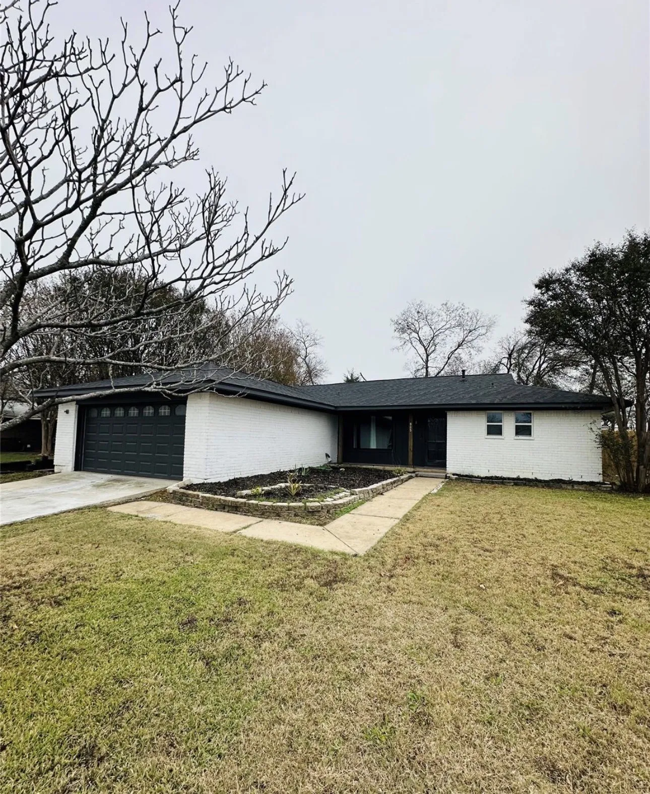 Single-story residence featuring white painted brick exterior, dark asphalt shingle roof, attached garage with sectional door, and raised stone-bordered garden bed