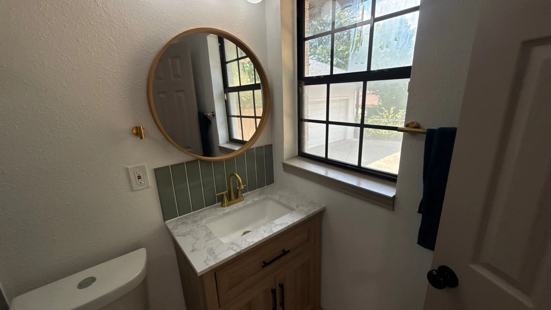 Wood-finish vanity with a stone-patterned countertop, integrated basin, vertical tile backsplash, round mirror, and brass-finish hardware