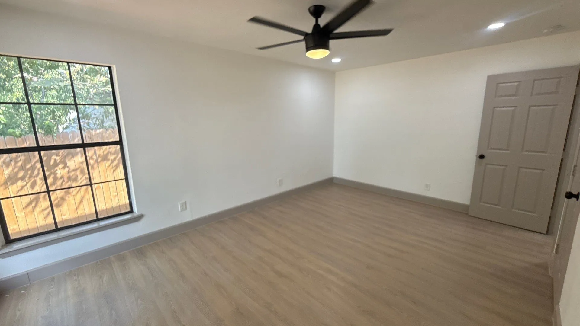 Interior room featuring wood-finish flooring, a multi-pane window with black framing, a matte black ceiling fan with an integrated light fixture, and a neutral-toned paneled door