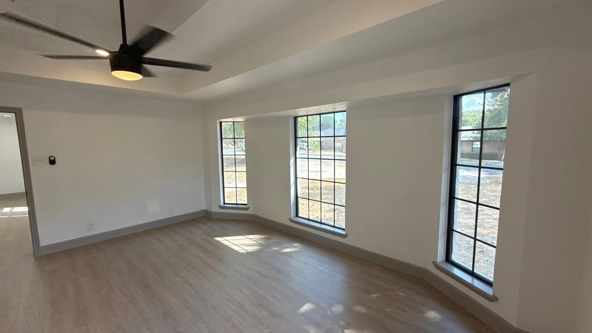 Tray ceiling with a matte black modern ceiling fan, wood-finish flooring, and expansive windows featuring dark frames