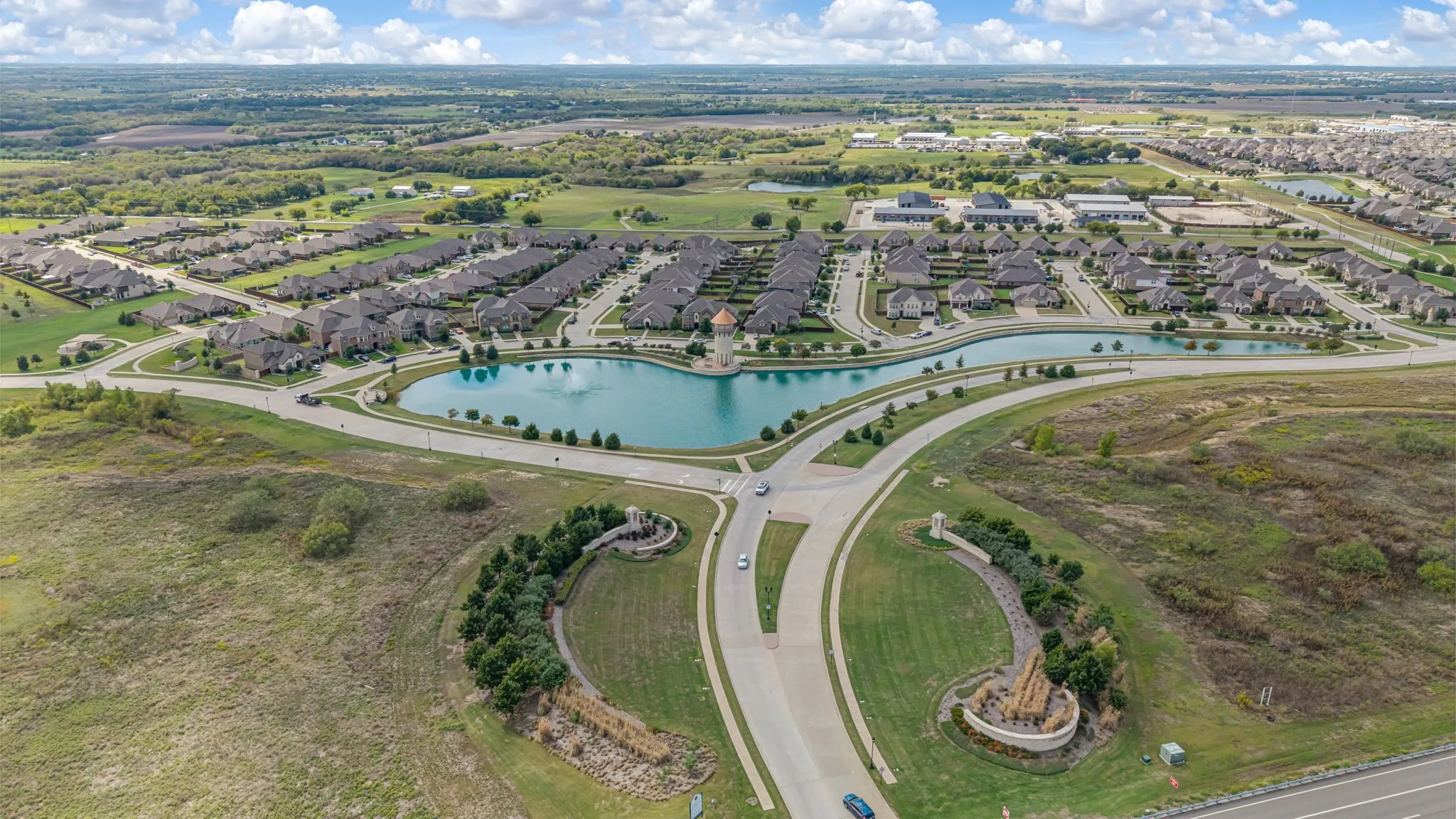 Aerial perspective of a residential community featuring a central pond with a fountain, landscaped entrance signage, and paved street infrastructure