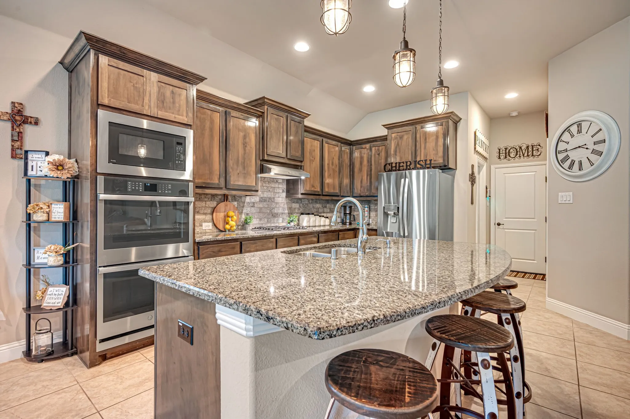 Kitchen featuring stained wood cabinetry, stone-finish countertops, stainless steel double wall ovens, a center island with integrated sink, and pendant lighting